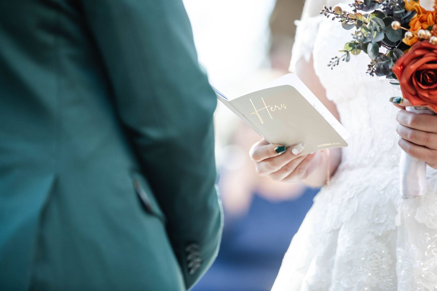 Bride reads vows from small booklet while holding red floral bouquet during indoor wedding ceremony.