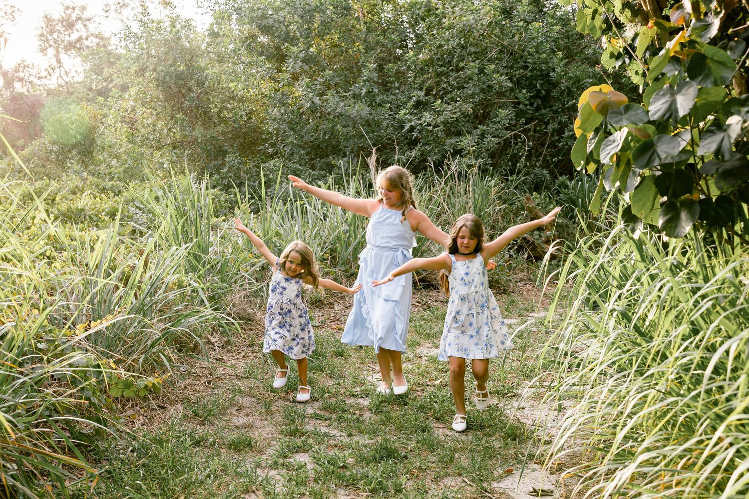 Three girls in floral dresses hold hands and dance along a sunlit nature path surrounded by lush green foliage.