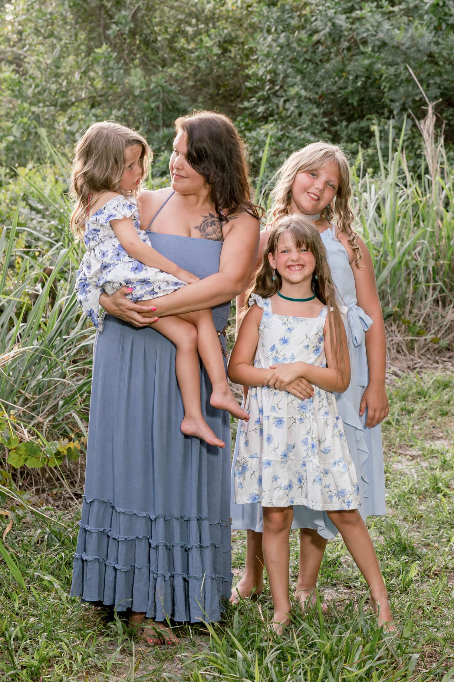 A mother in a blue maxi dress holds a young girl while two other girls pose outdoors in a lush green setting.