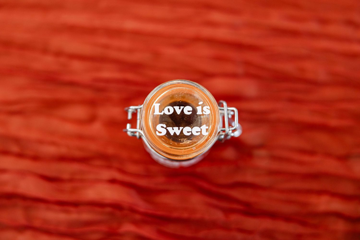 Top view of glass jar filled with honey labeled Love is Sweet on red fabric.