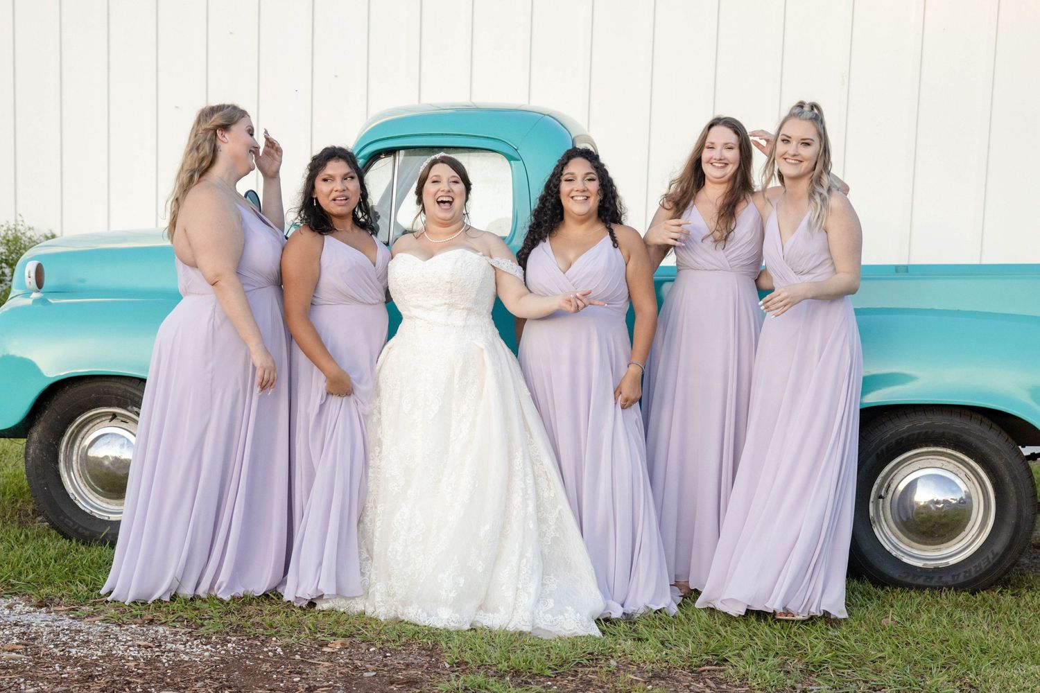 Bride with bridesmaids in lavender gowns laughing beside a vintage turquoise truck outdoors.