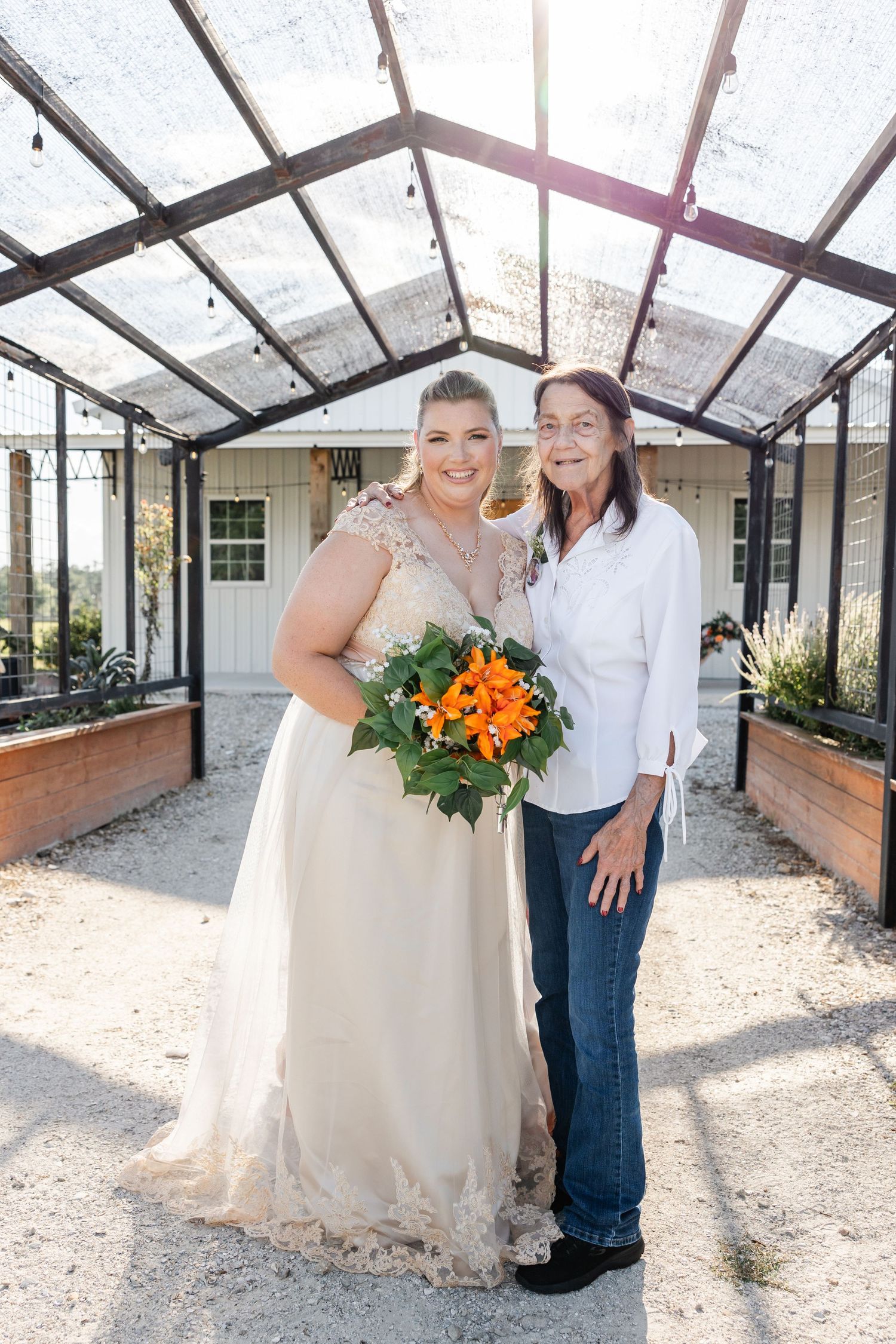 Bride in ivory lace gown poses with older woman holding orange bouquet under greenhouse structure.