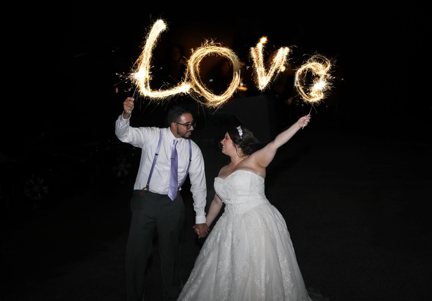 Bride and groom holding hands while sparklers write Love in the night sky at their wedding celebration.