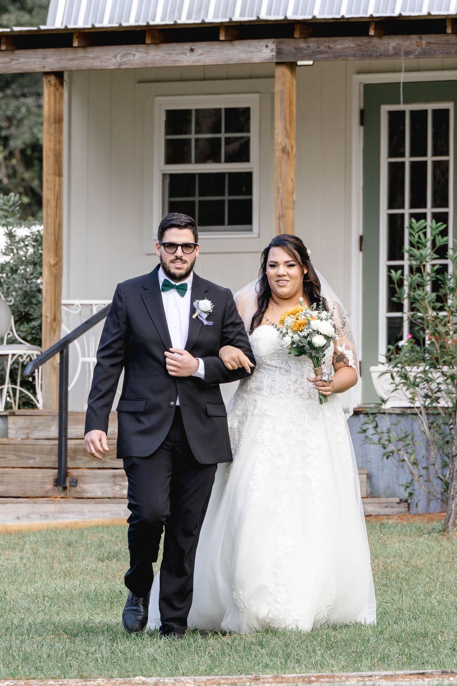 Bride in lace gown holding sunflower bouquet walks with groom in black tuxedo and green bow tie.