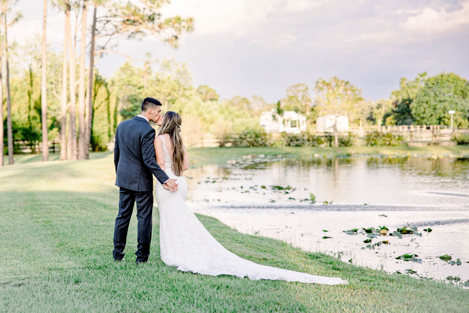 Bride and groom kissing near a serene pond with a long flowing white wedding dress train.