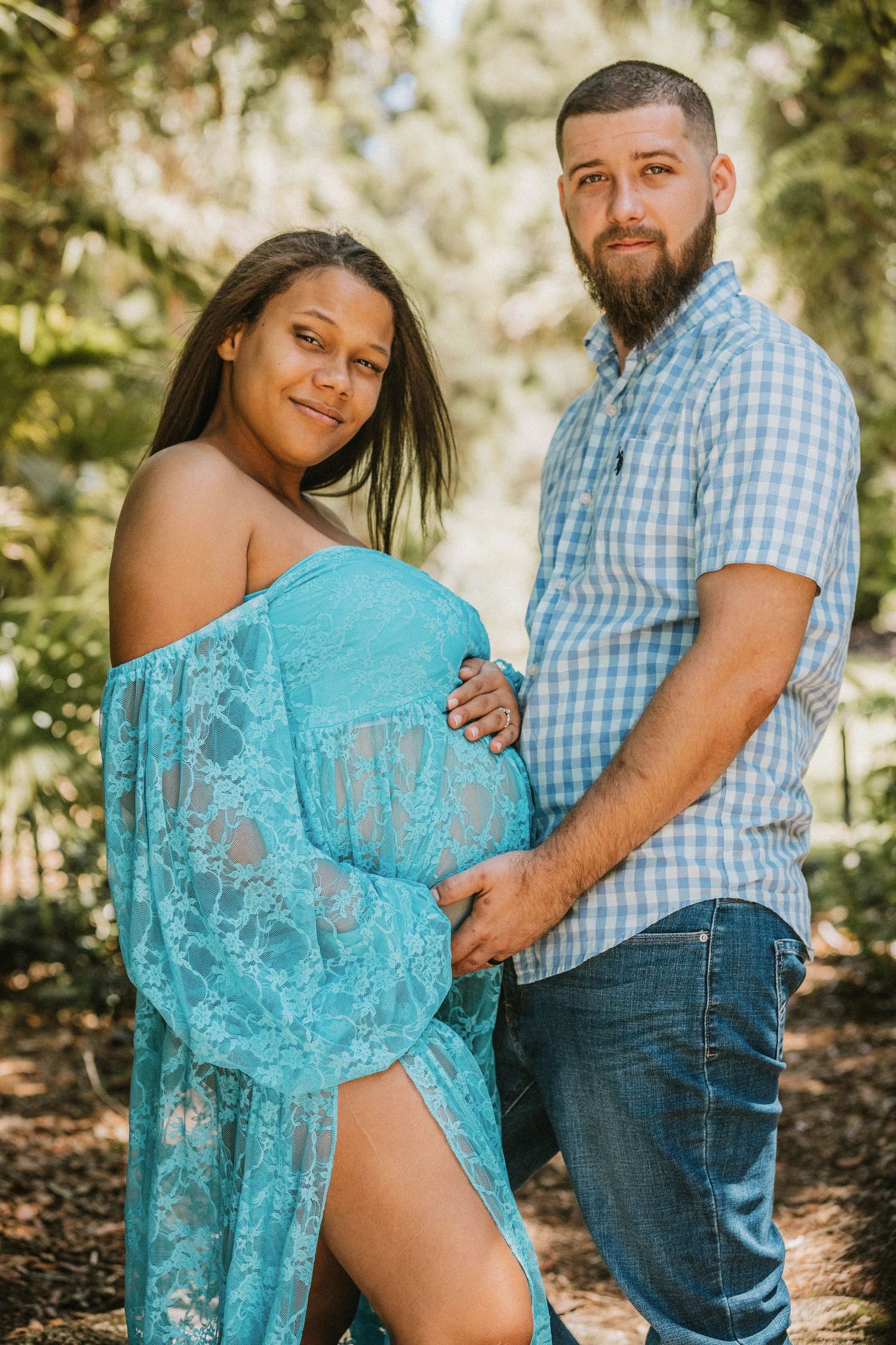 Pregnant woman in teal lace maternity gown poses with bearded man outdoors in a lush green forest setting.