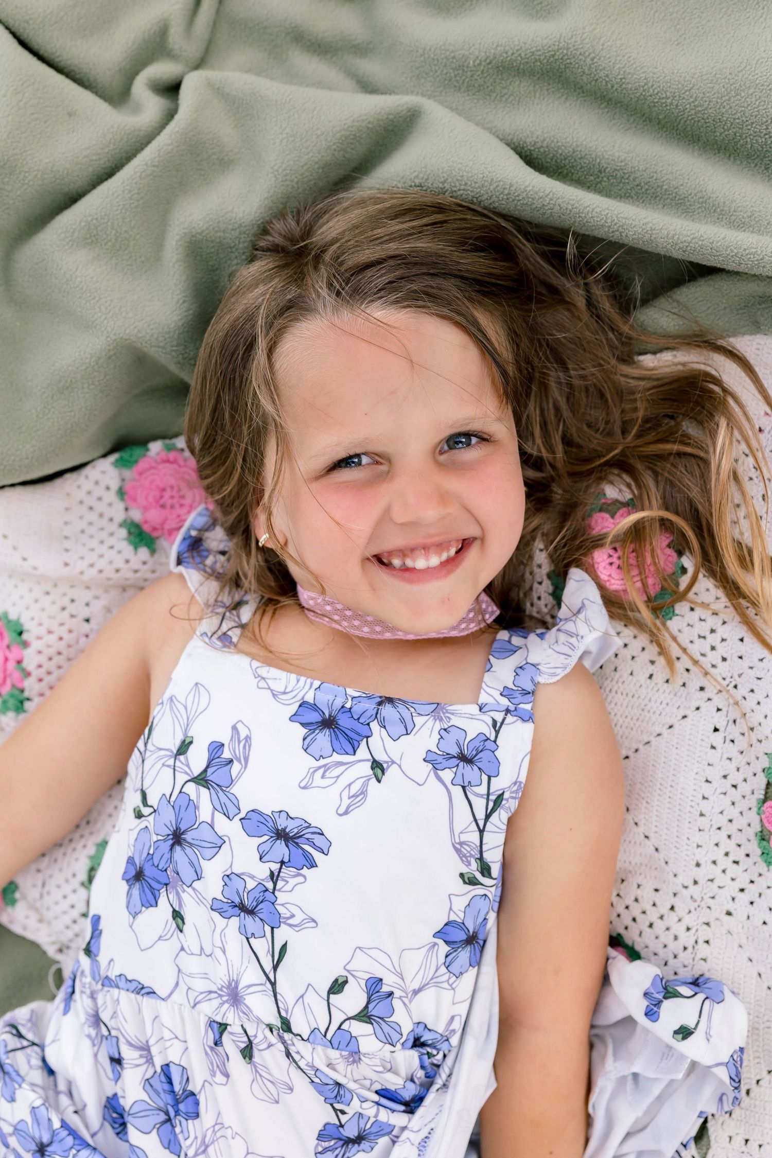 Smiling young girl in a blue floral dress lying on a blanket, looking up at the camera with bright blue eyes.