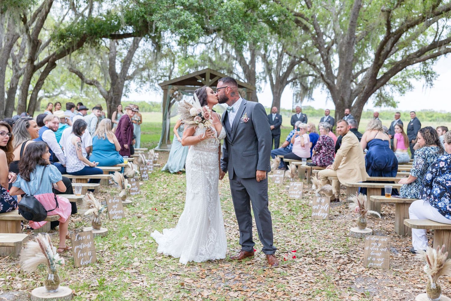 Newlyweds share first kiss at outdoor wedding ceremony under oak trees with seated guests.
