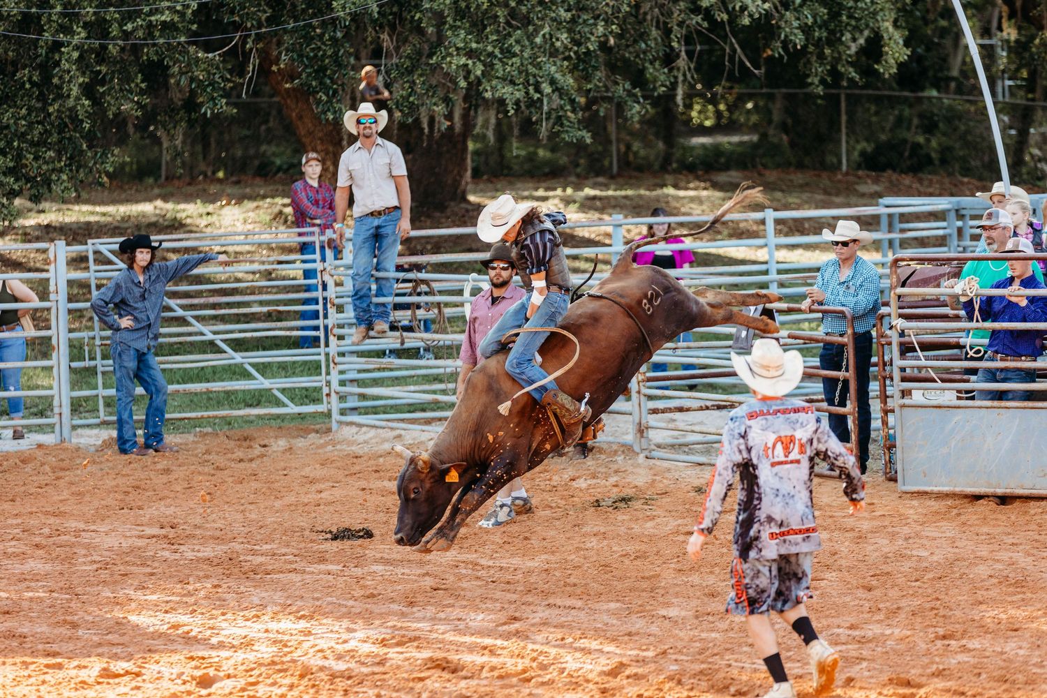 Bull rider competing in rodeo arena while clown stands nearby during bucking bull event.