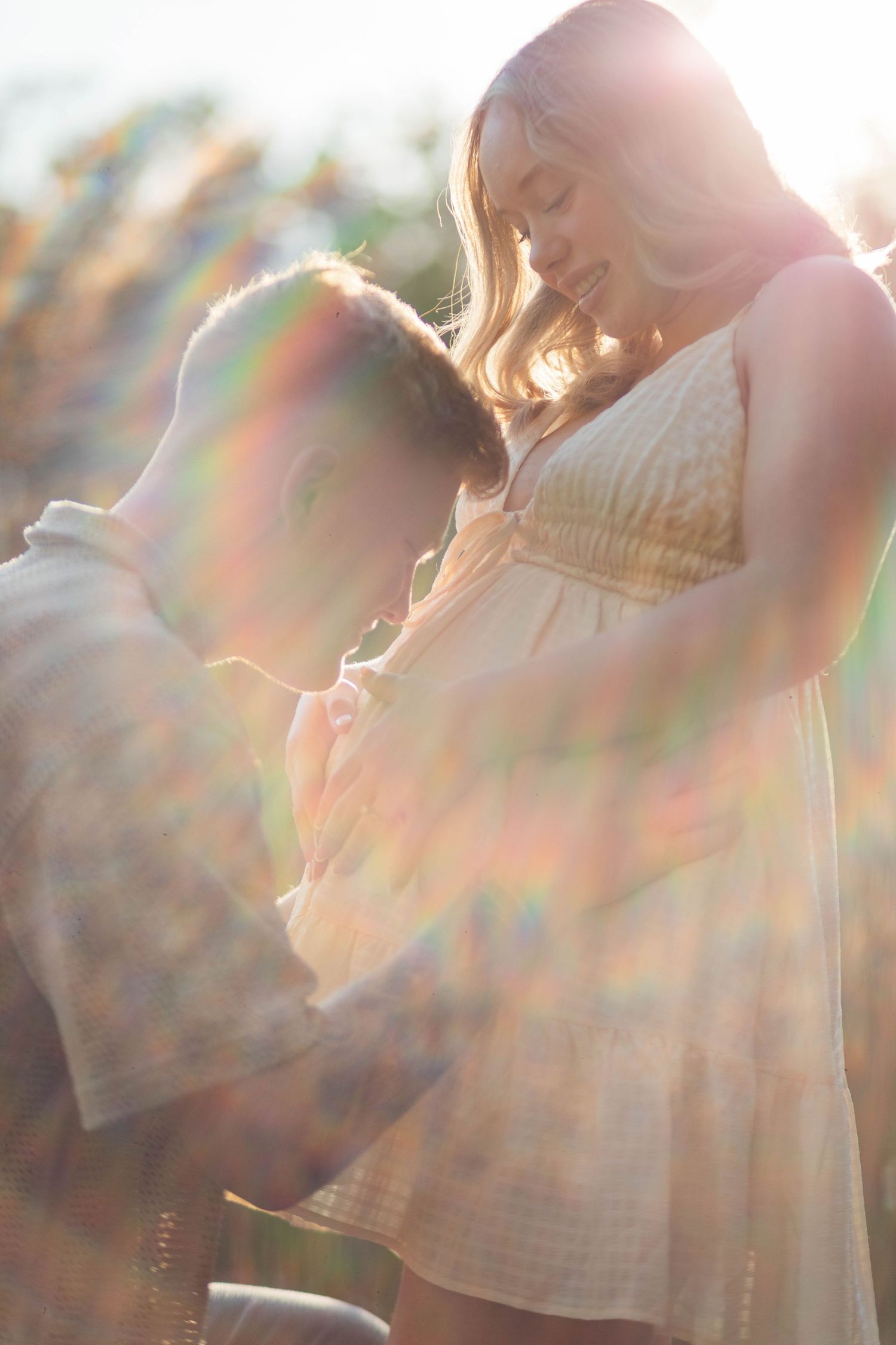 Couple holding hands in golden sunlight with rainbow lens flare during outdoor maternity photo session.