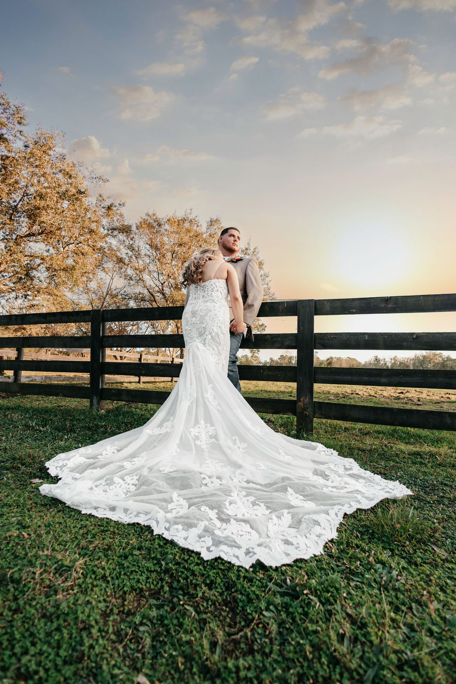 Bride and groom at sunset by wooden fence with stunning lace mermaid wedding gown train.