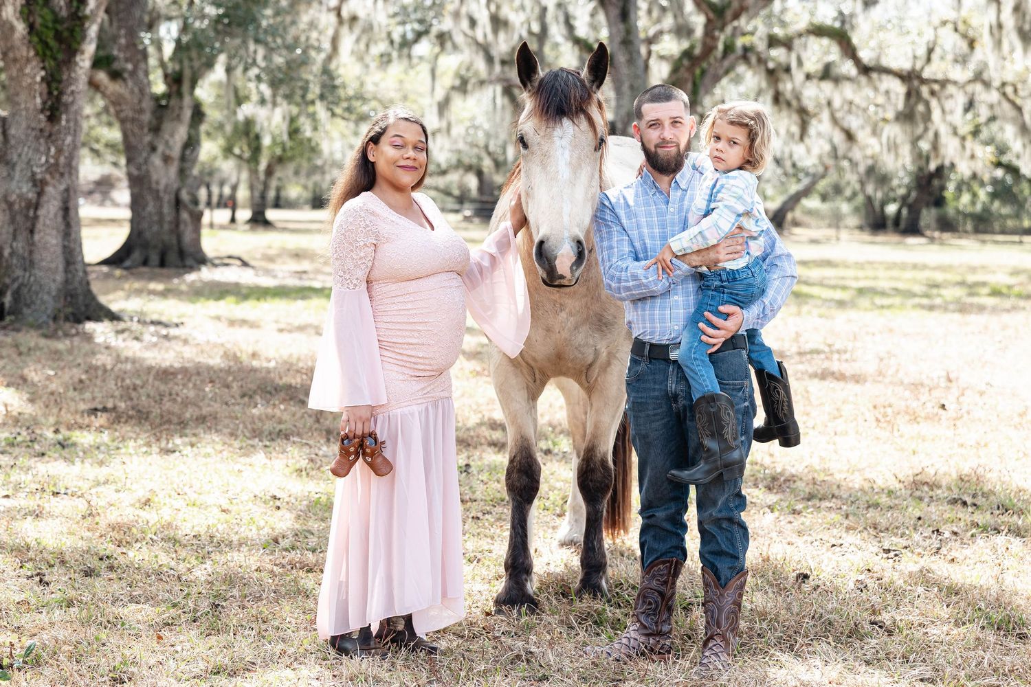 Pregnant woman in pink gown and man holding toddler pose with a gray horse outdoors under moss-covered trees.
