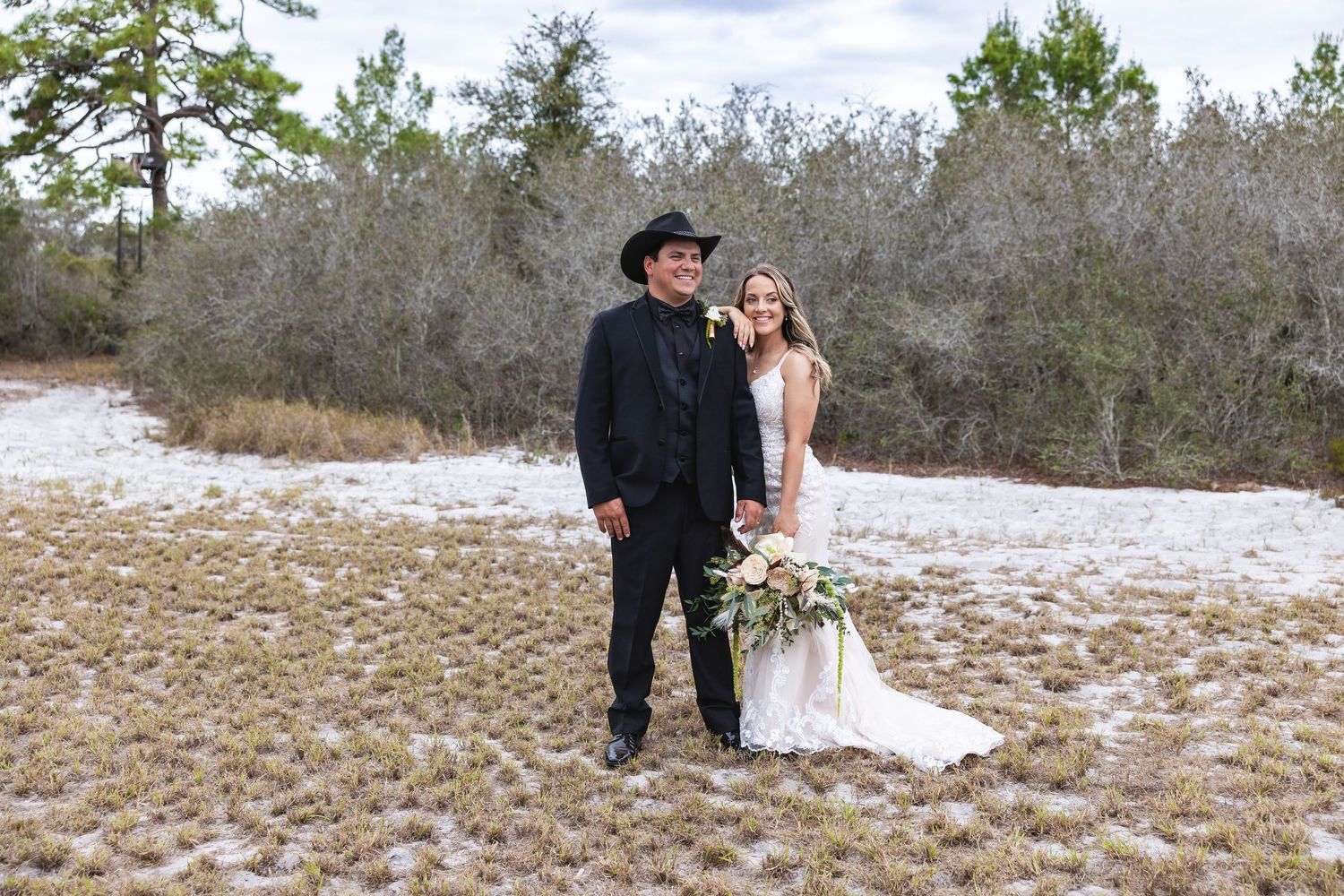 Bride and groom posing together in a rustic outdoor setting, groom wearing a black cowboy hat and suit.