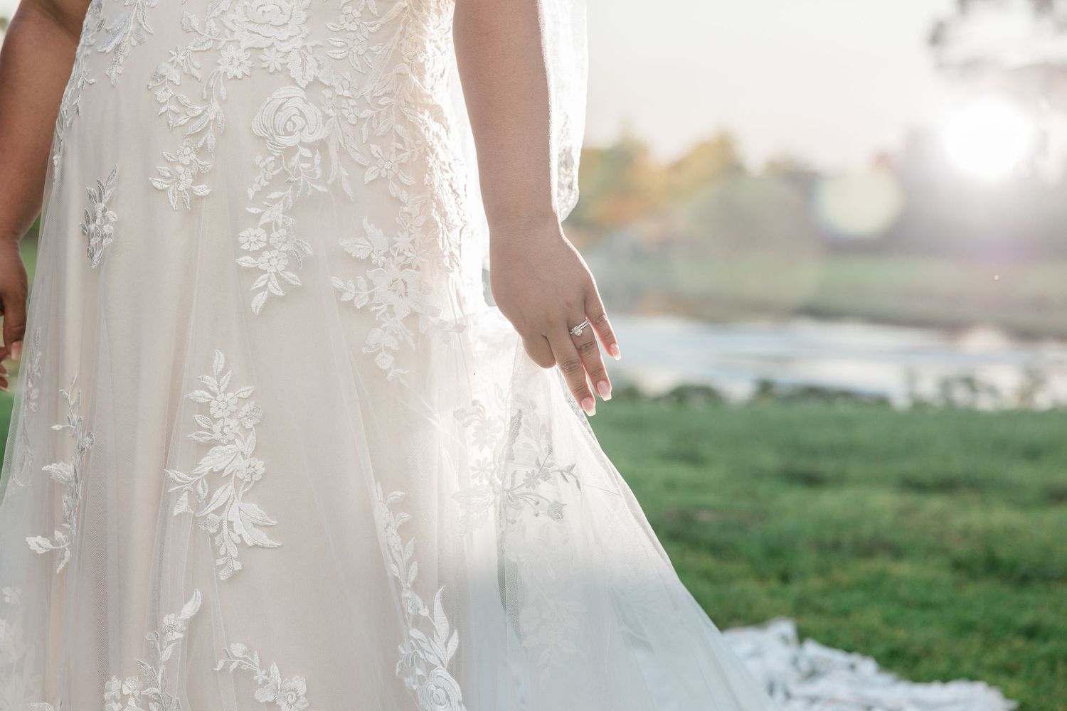 Close-up detail of a bride in a floral lace wedding gown with a sparkling ring, standing outdoors.