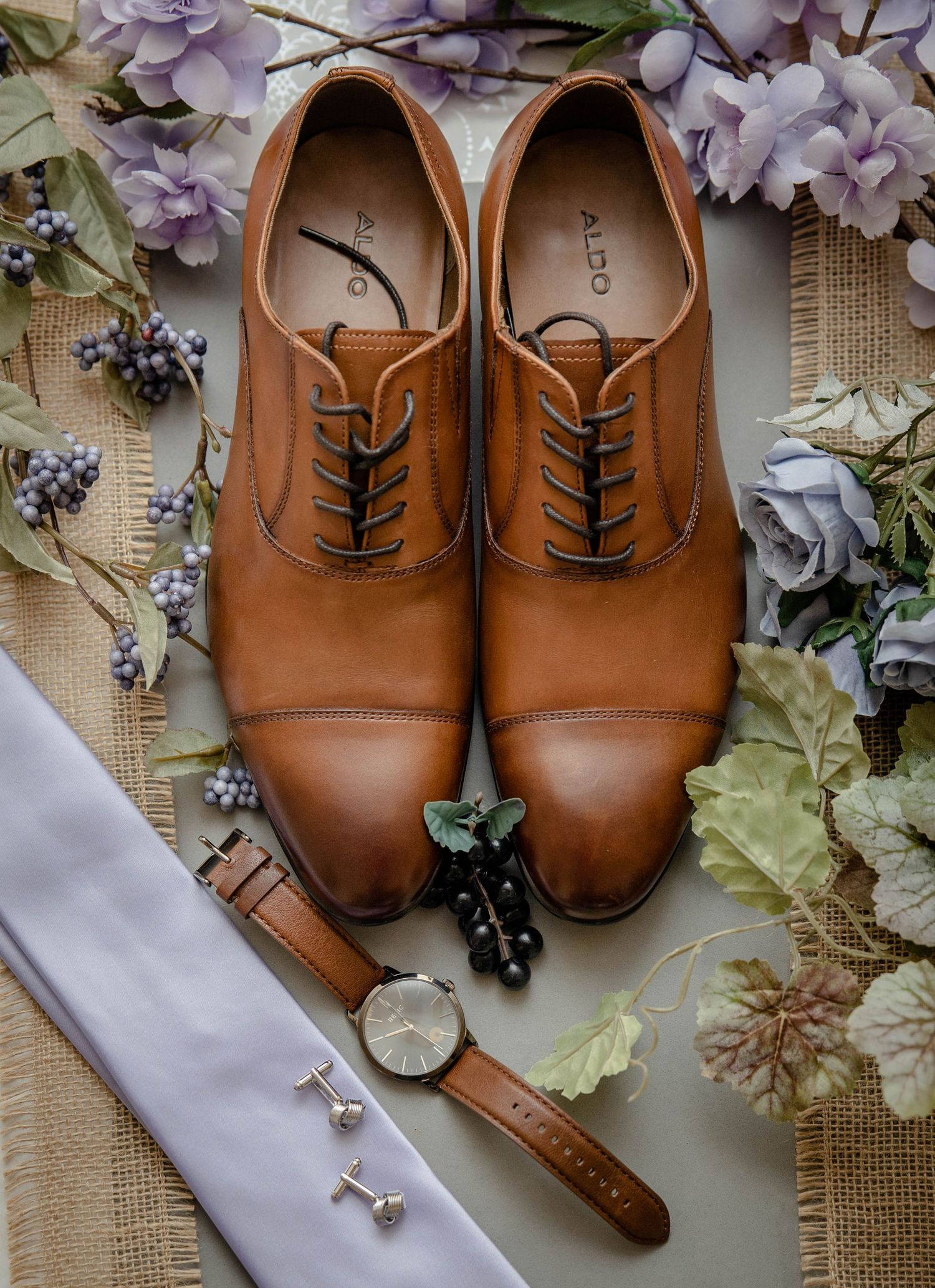 Aldo brown leather Oxford shoes with watch, cufflinks and floral boutonniere wedding accessories flatlay.