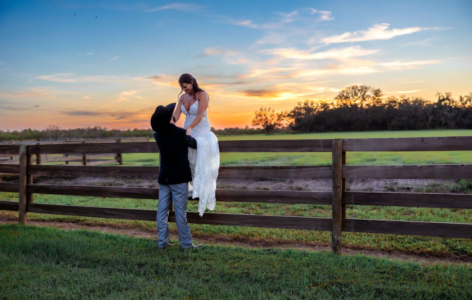 Groom lifts bride in white wedding gown at sunset by a wooden farm fence in a romantic outdoor portrait.