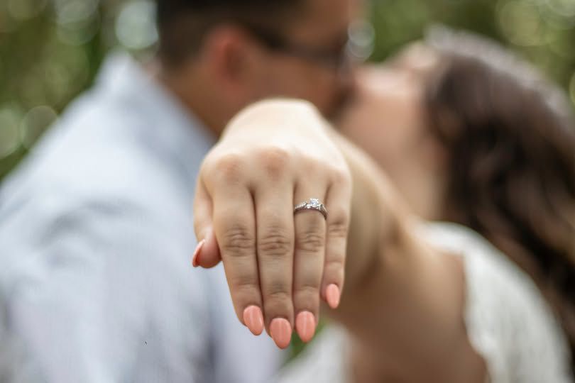 Couple kissing while showing off diamond engagement ring with pink manicure outdoors.