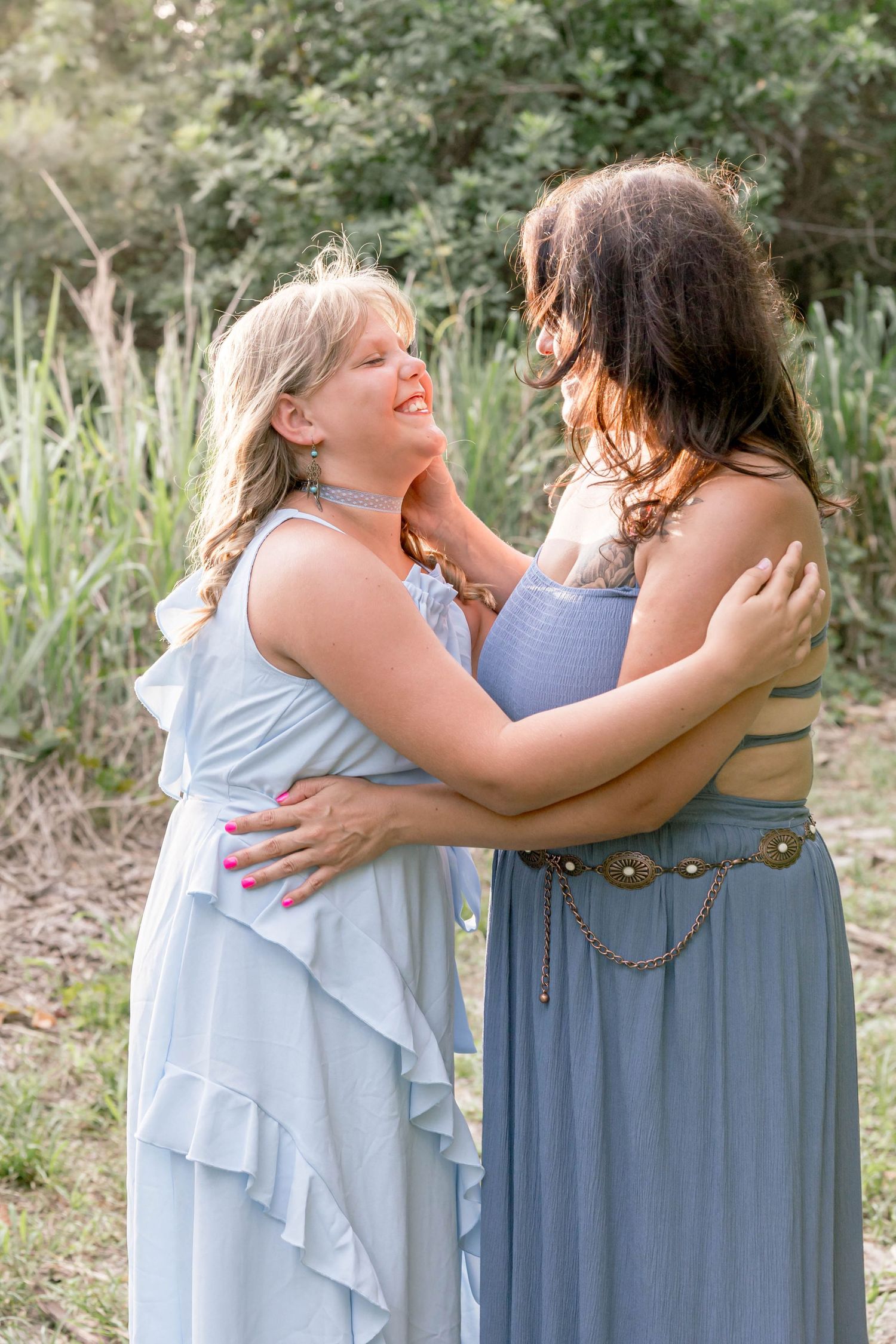 Two women in blue dresses embracing warmly in a lush green outdoor setting.