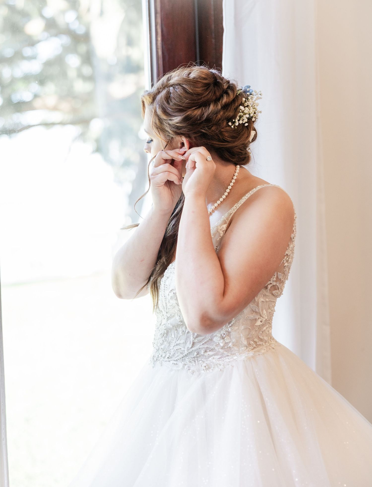 Bride in beaded ballgown with floral updo adjusting earring by window in soft natural light.