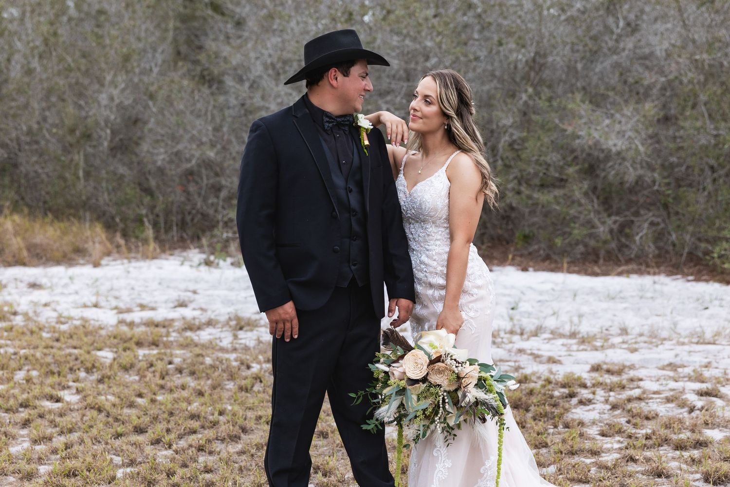 Bride holding bouquet gazes at groom in black cowboy hat against snowy winter landscape.