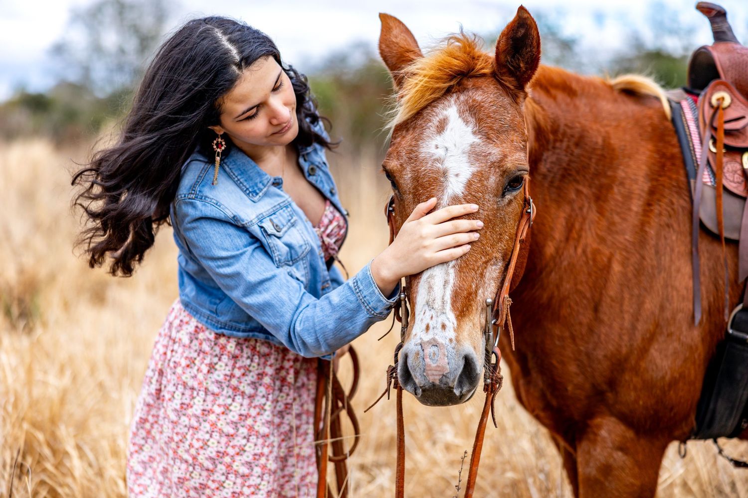 Young woman in denim jacket petting chestnut horse in golden dry grass field.