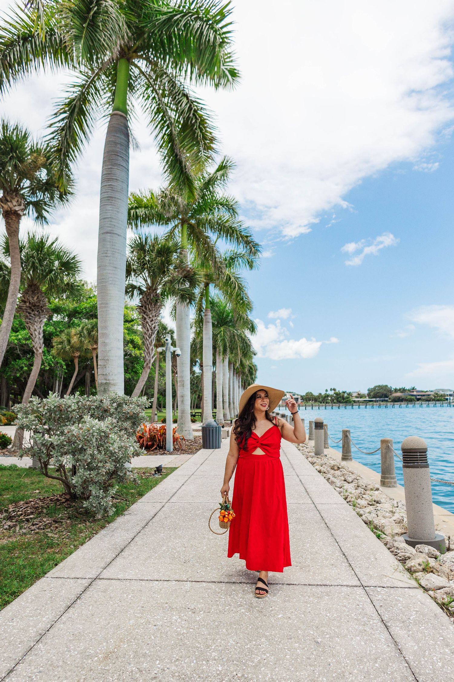 Woman in red maxi dress and straw hat strolling along palm tree-lined waterfront promenade on sunny day.
