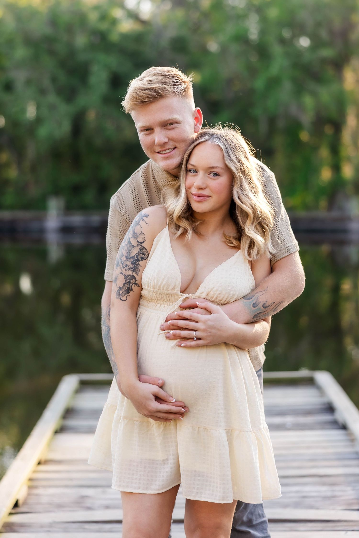 Smiling tattooed couple embracing on a wooden dock, cradling a baby bump in warm golden light.