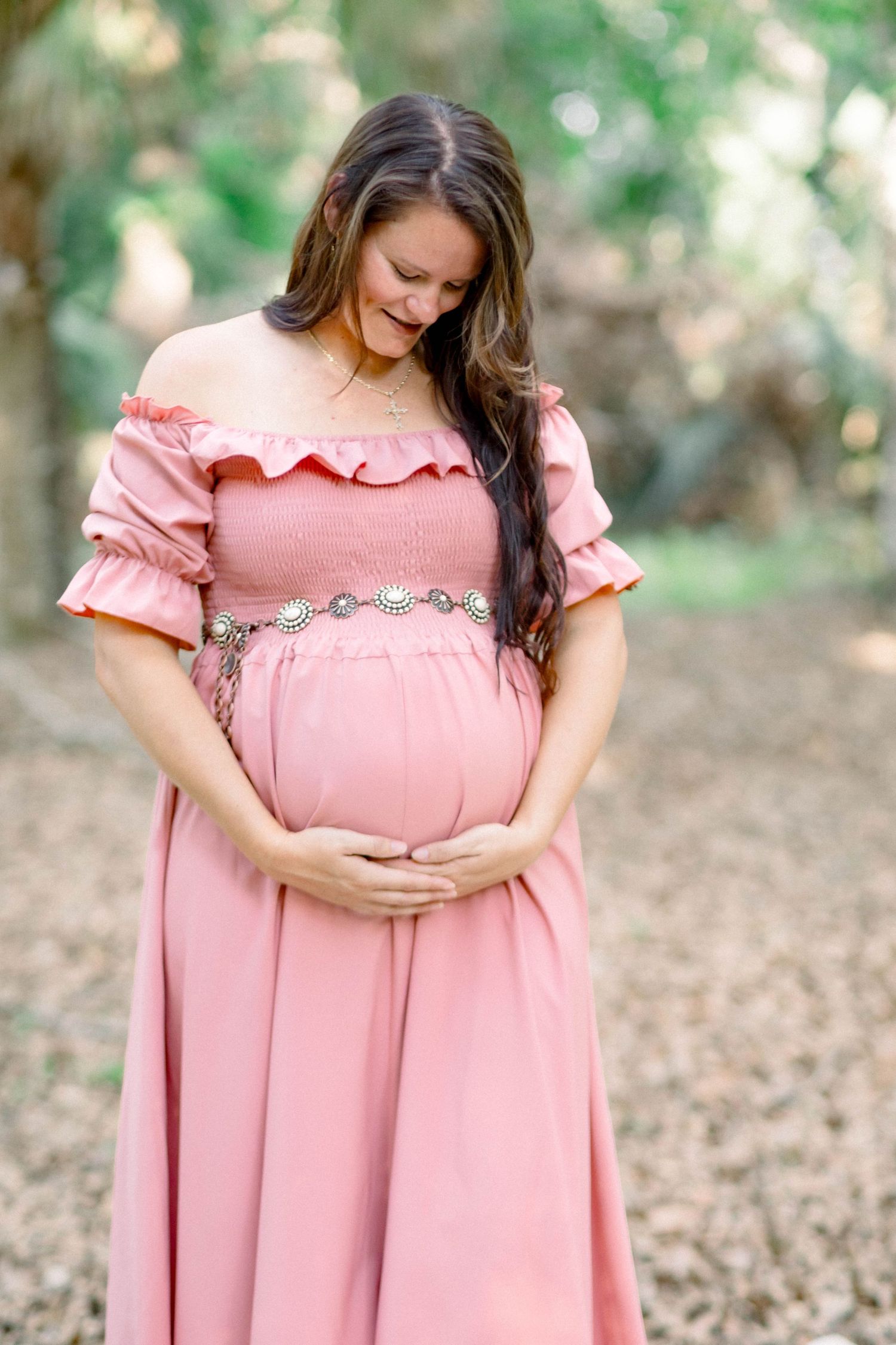 Expectant mother in blush pink off-shoulder dress with silver belt cradling baby bump outdoors.