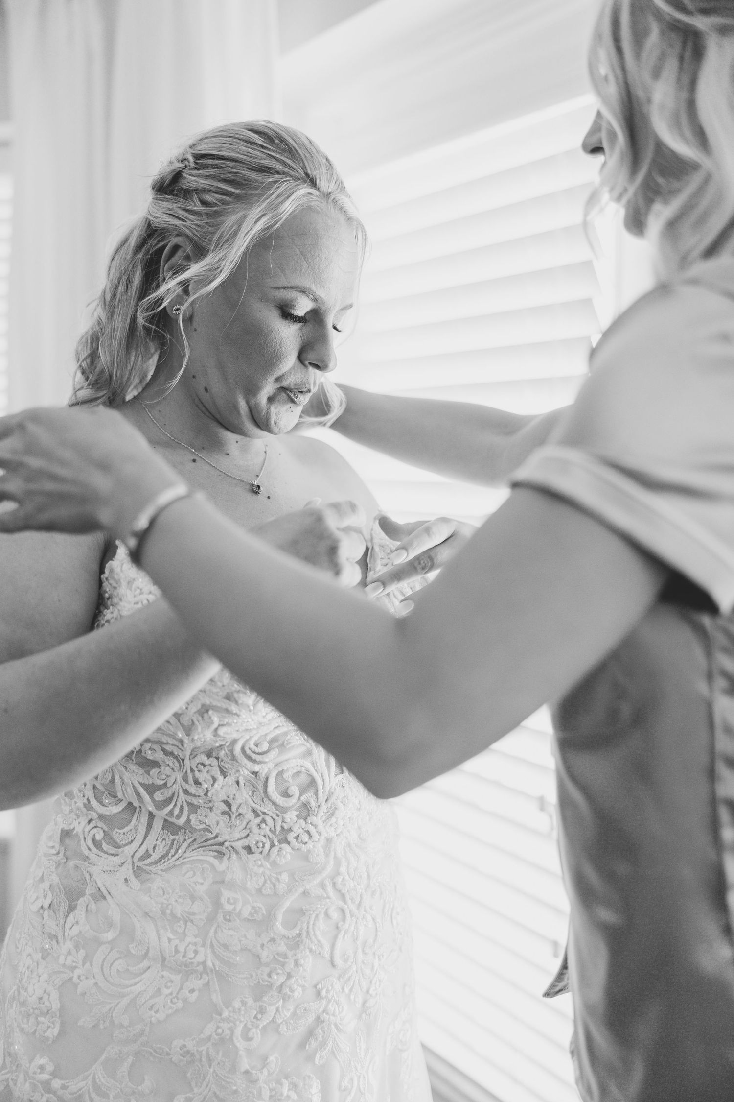 Black and white photo of bride in lace gown being helped to dress near a bright window.