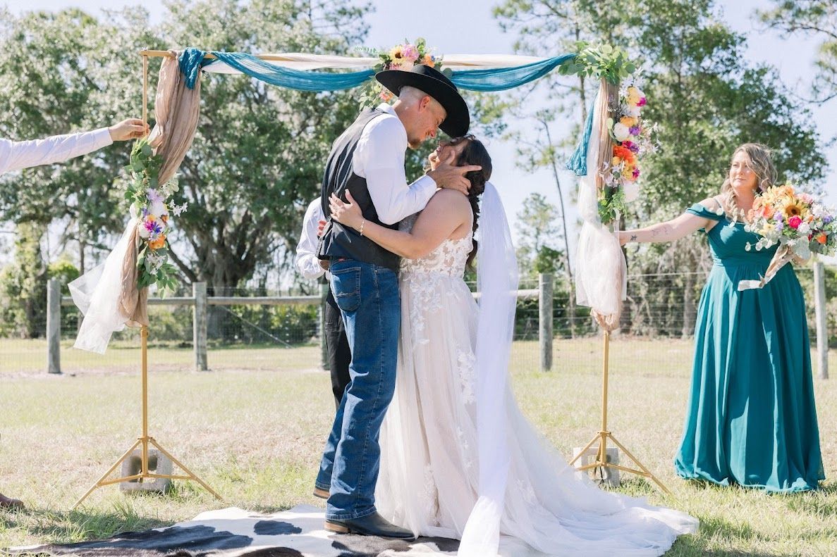 Bride and groom share first kiss under colorful floral arch at outdoor rustic wedding ceremony.