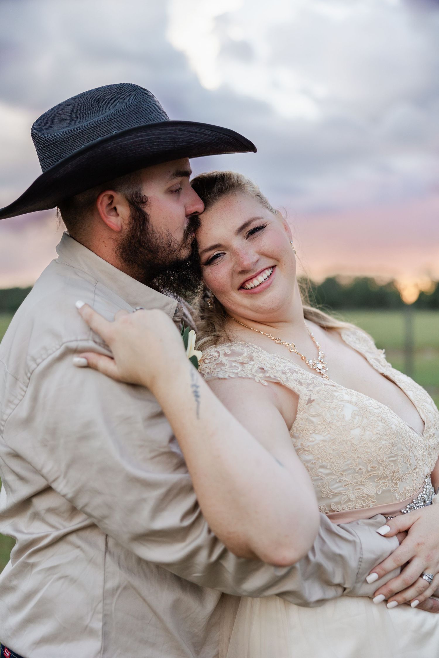 Groom in cowboy hat kisses bride's cheek as she smiles during outdoor sunset wedding portrait.