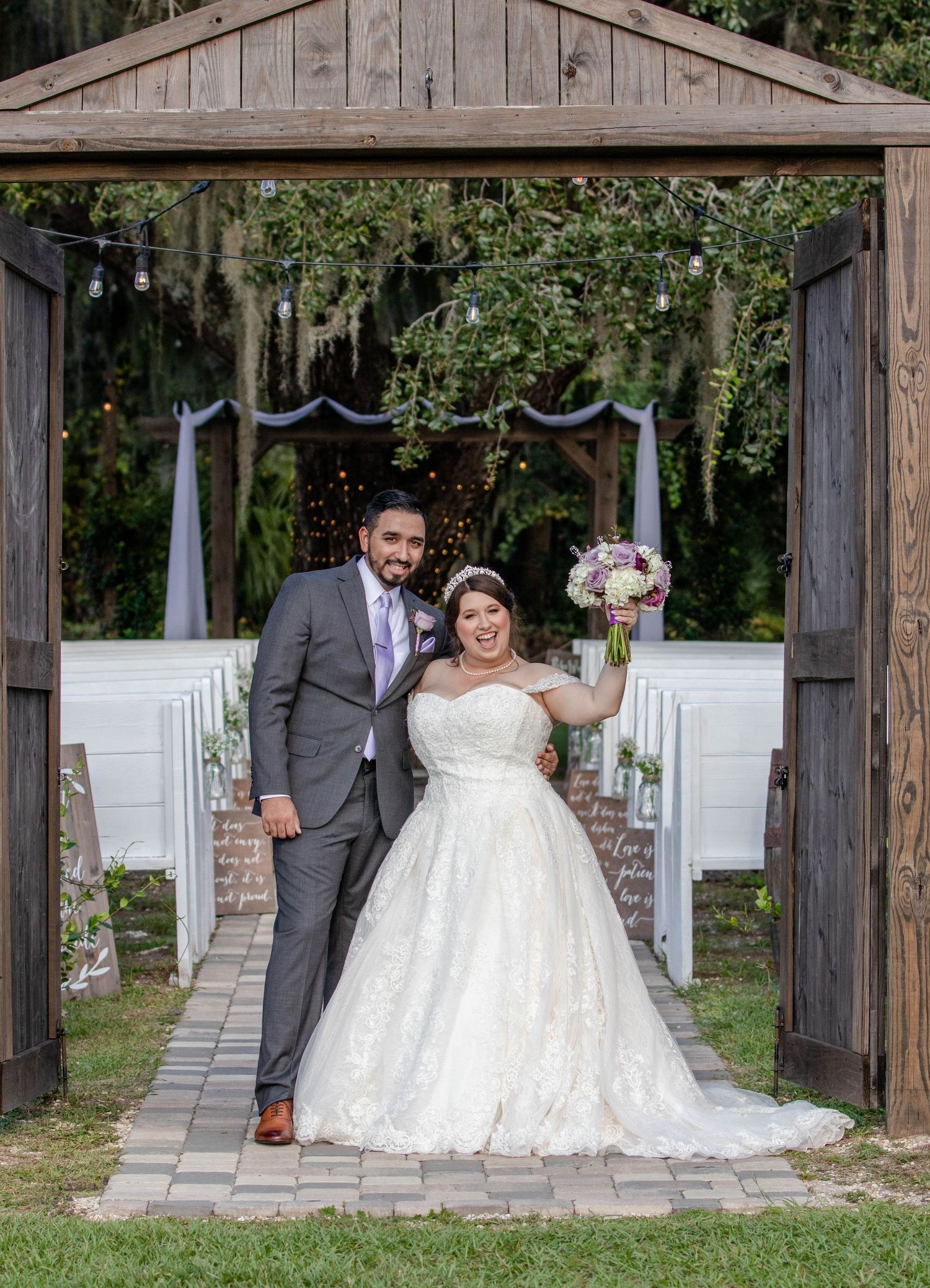 Newlywed couple posing under rustic wooden arch at outdoor wedding ceremony venue with string lights.
