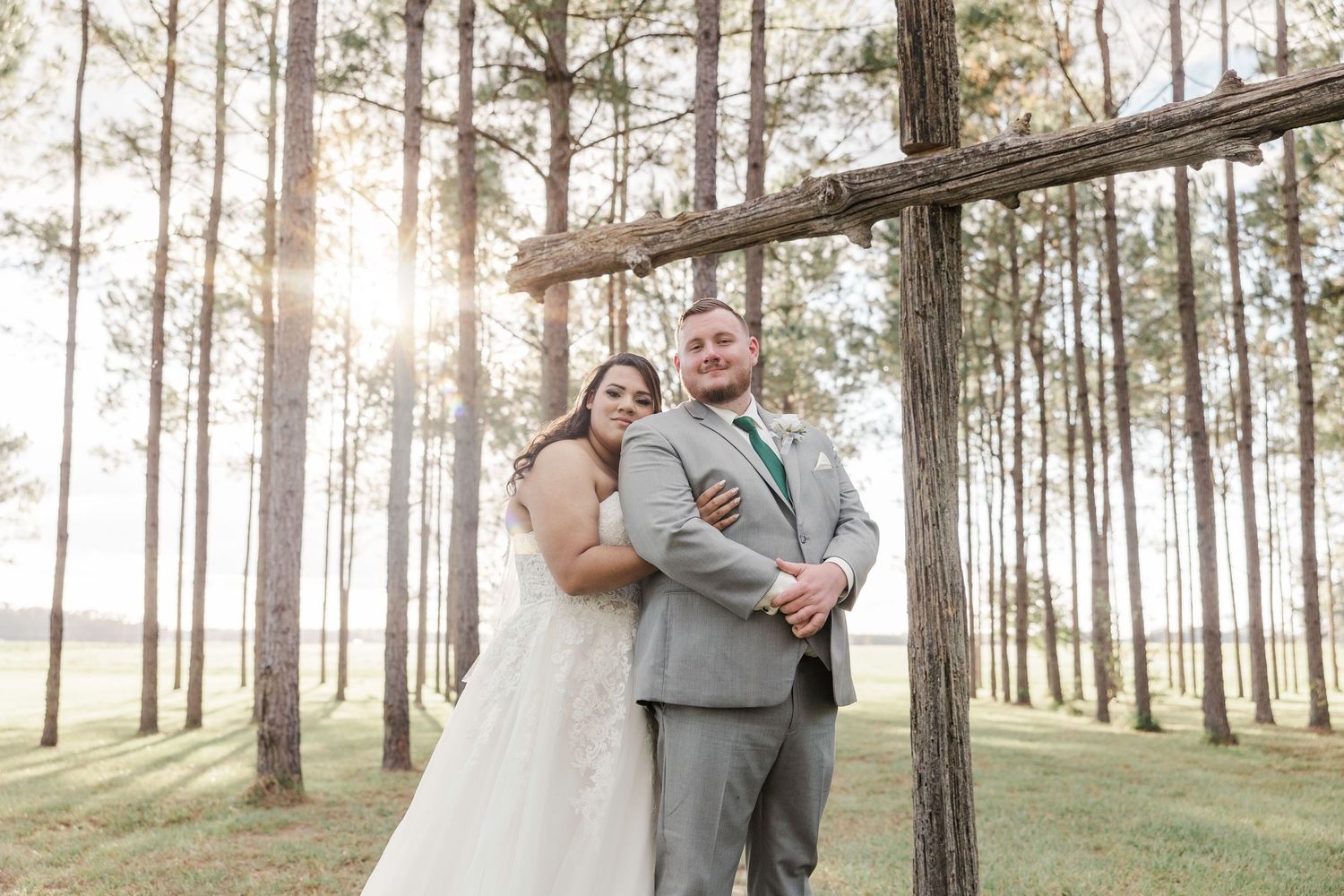 Bride and groom embrace near rustic wooden arch with sunlight streaming through tall pine trees.