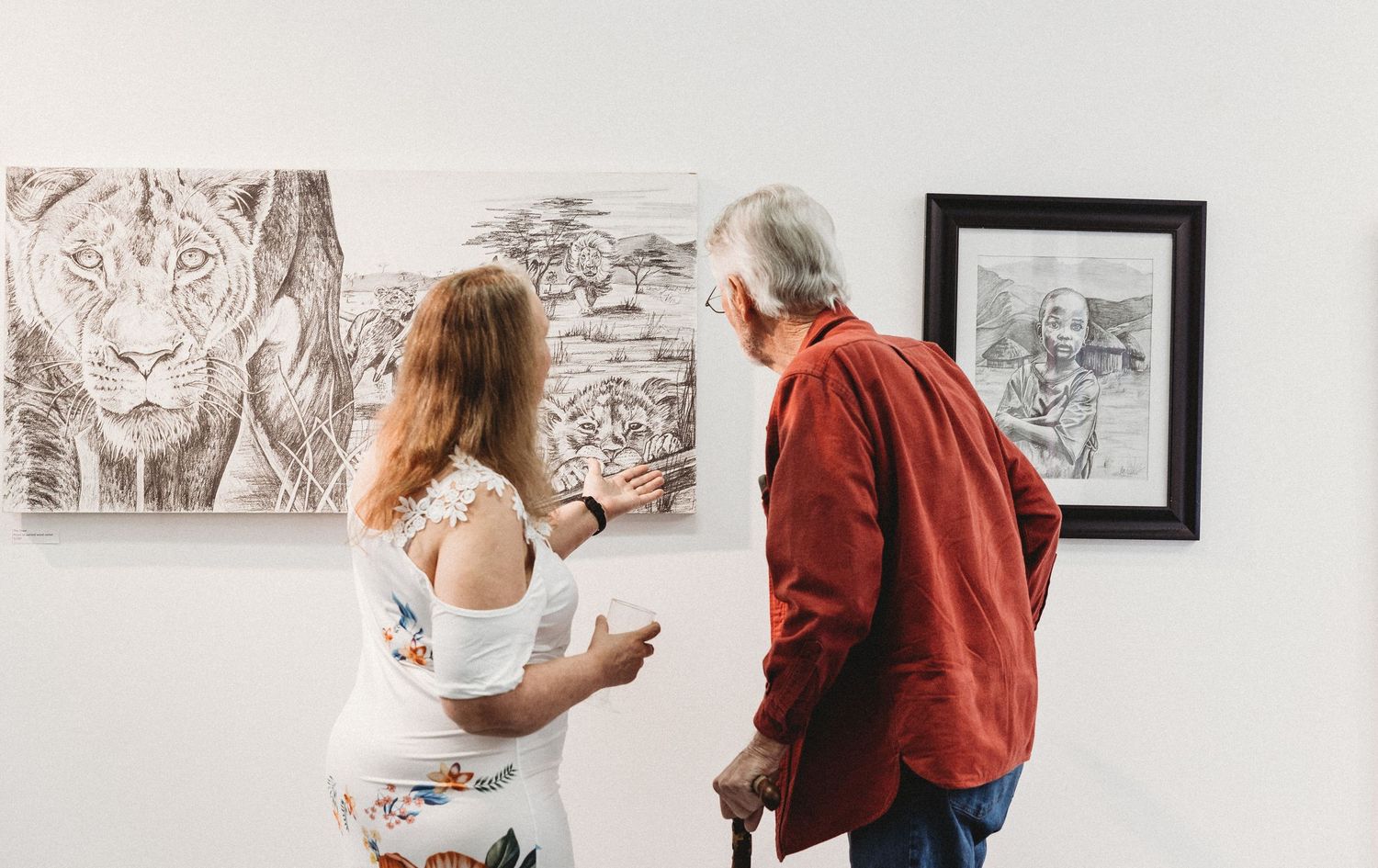 Woman and older man viewing large wildlife drawings including lion artwork at gallery exhibition.