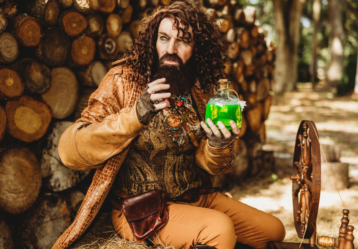 Man in medieval fantasy costume holding a glowing green potion bottle outdoors near stacked logs.