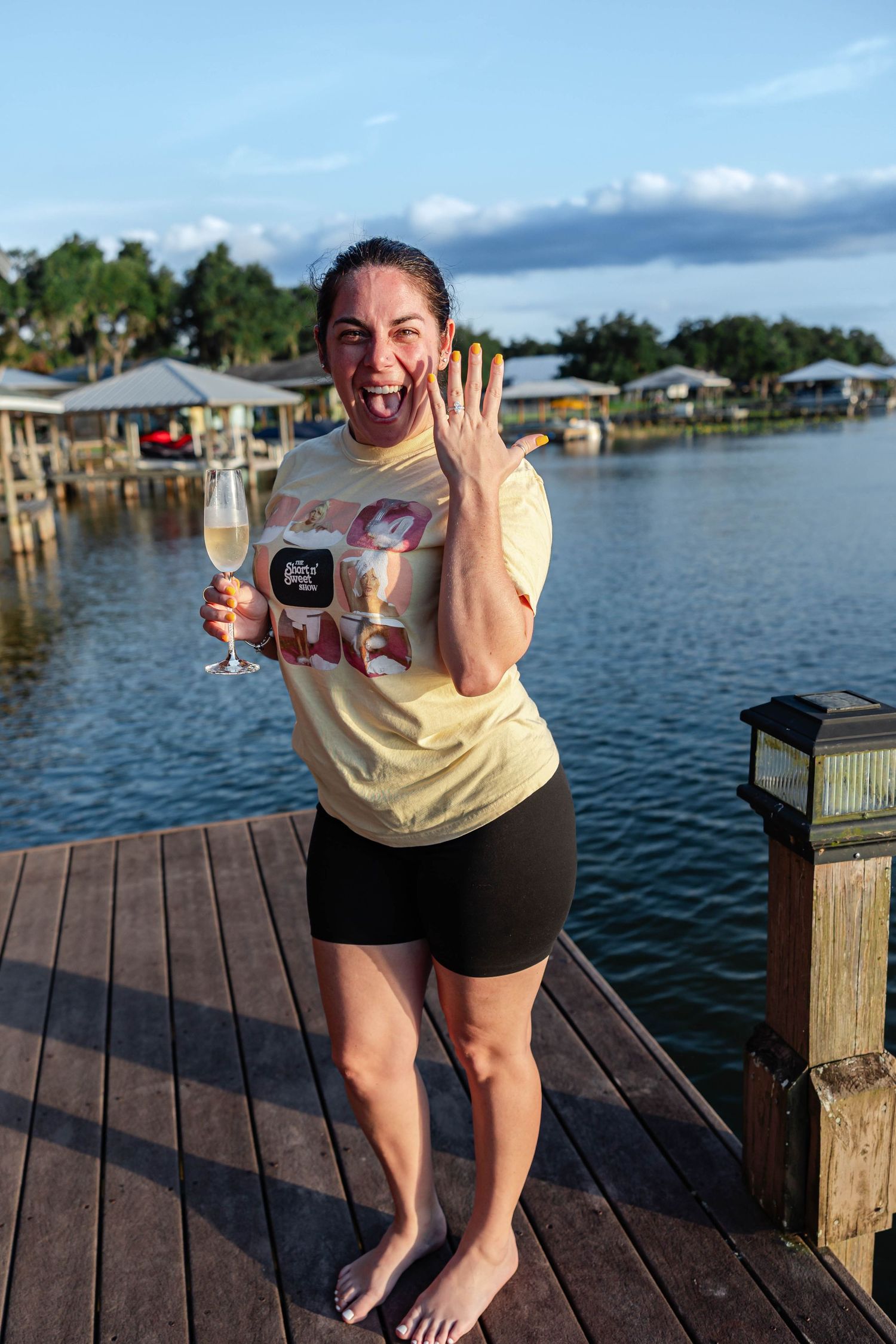 Excited woman showing engagement ring on dock, holding champagne glass, waterfront backdrop.