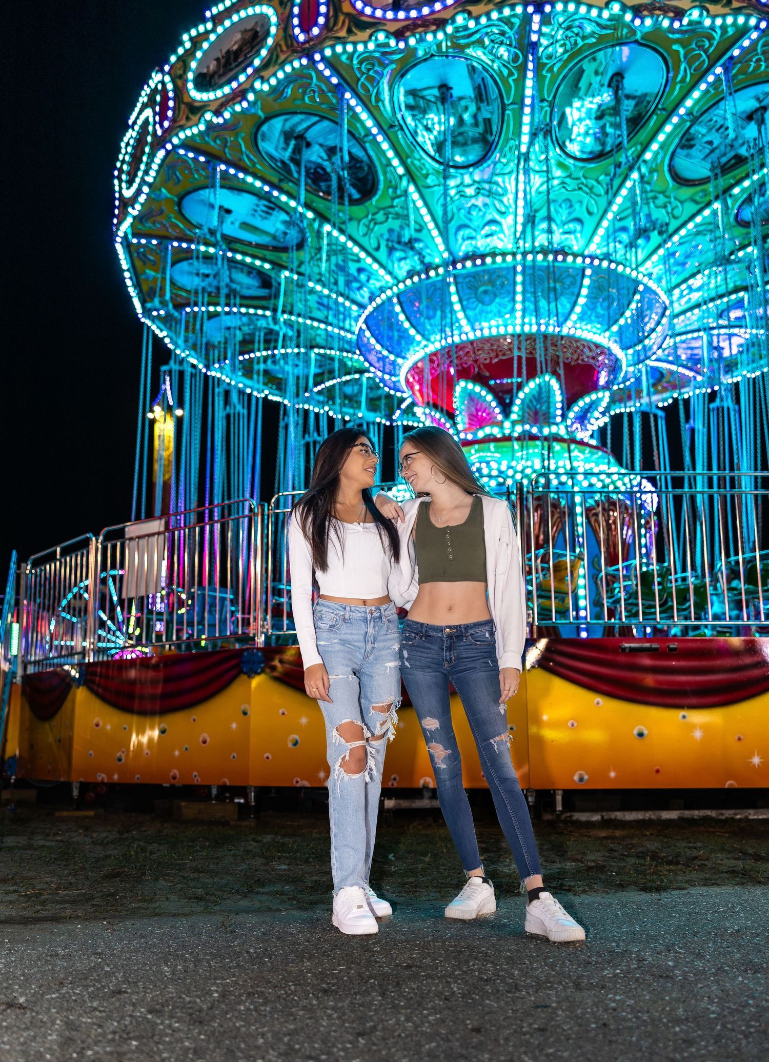 Two young women pose together in front of a glowing illuminated carousel at a nighttime carnival.