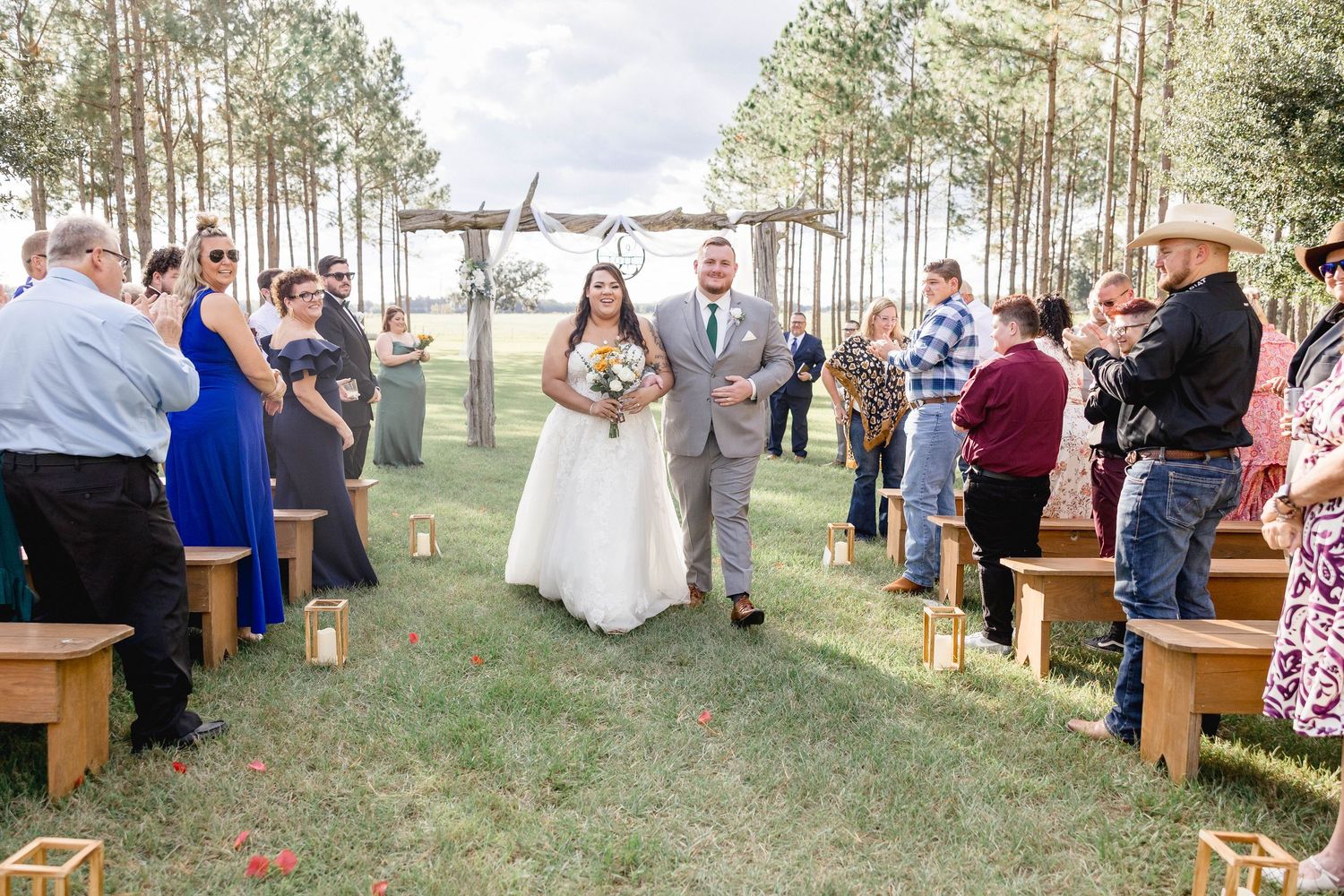 Bride and groom walk down outdoor aisle lined with wooden benches and candles surrounded by pine trees.