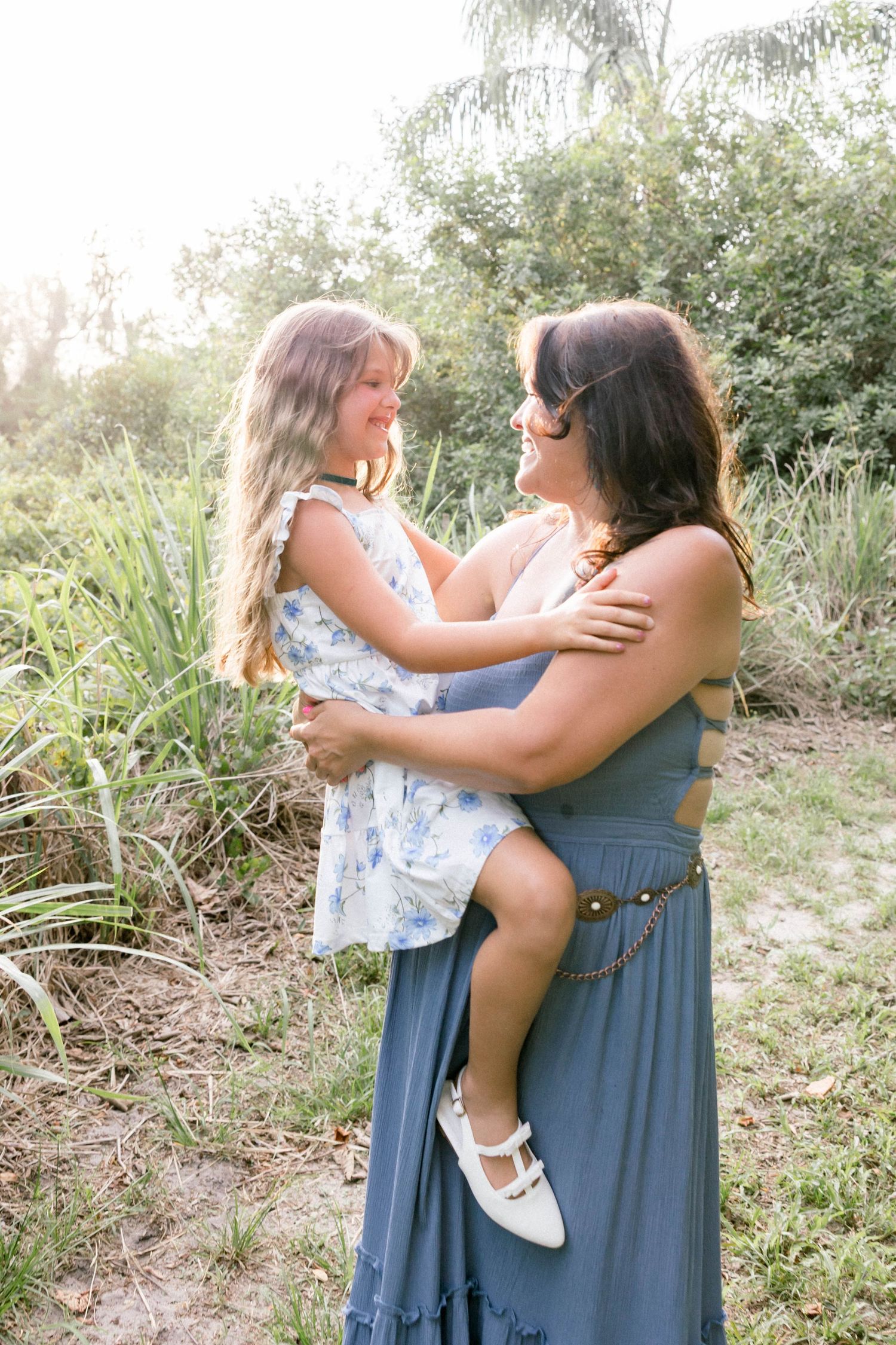 A woman in a blue dress holds a young girl in a floral dress outdoors among lush greenery.