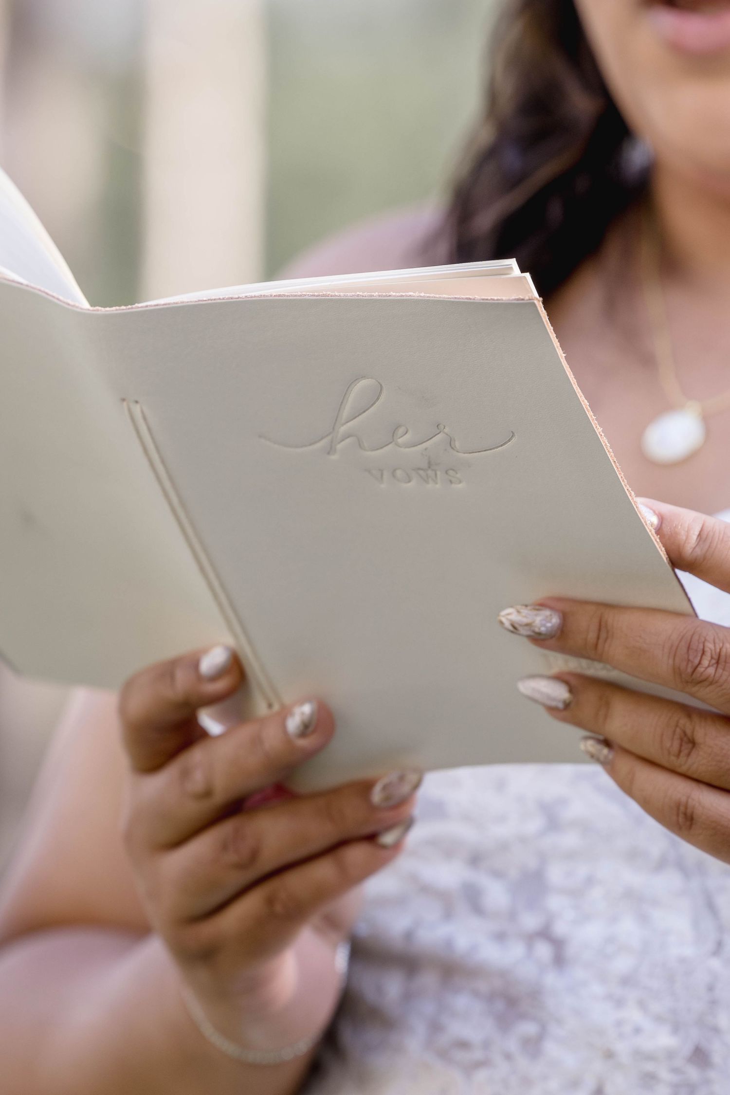 Bride reads wedding vows from a white embossed vow book during outdoor wedding ceremony.