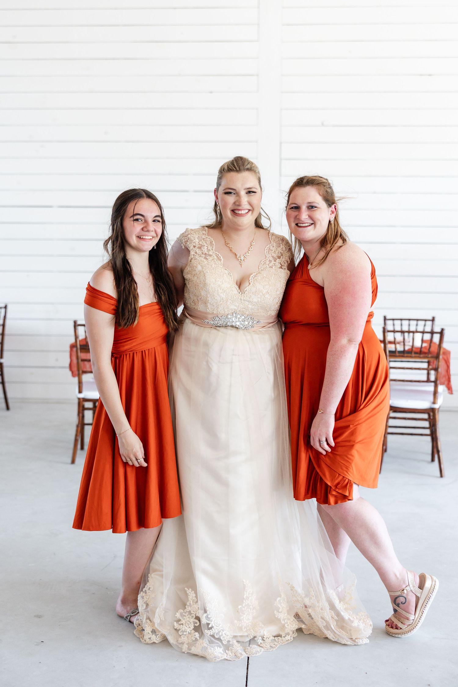 Bride in ivory lace gown stands with two bridesmaids in rust orange dresses inside white barn venue.