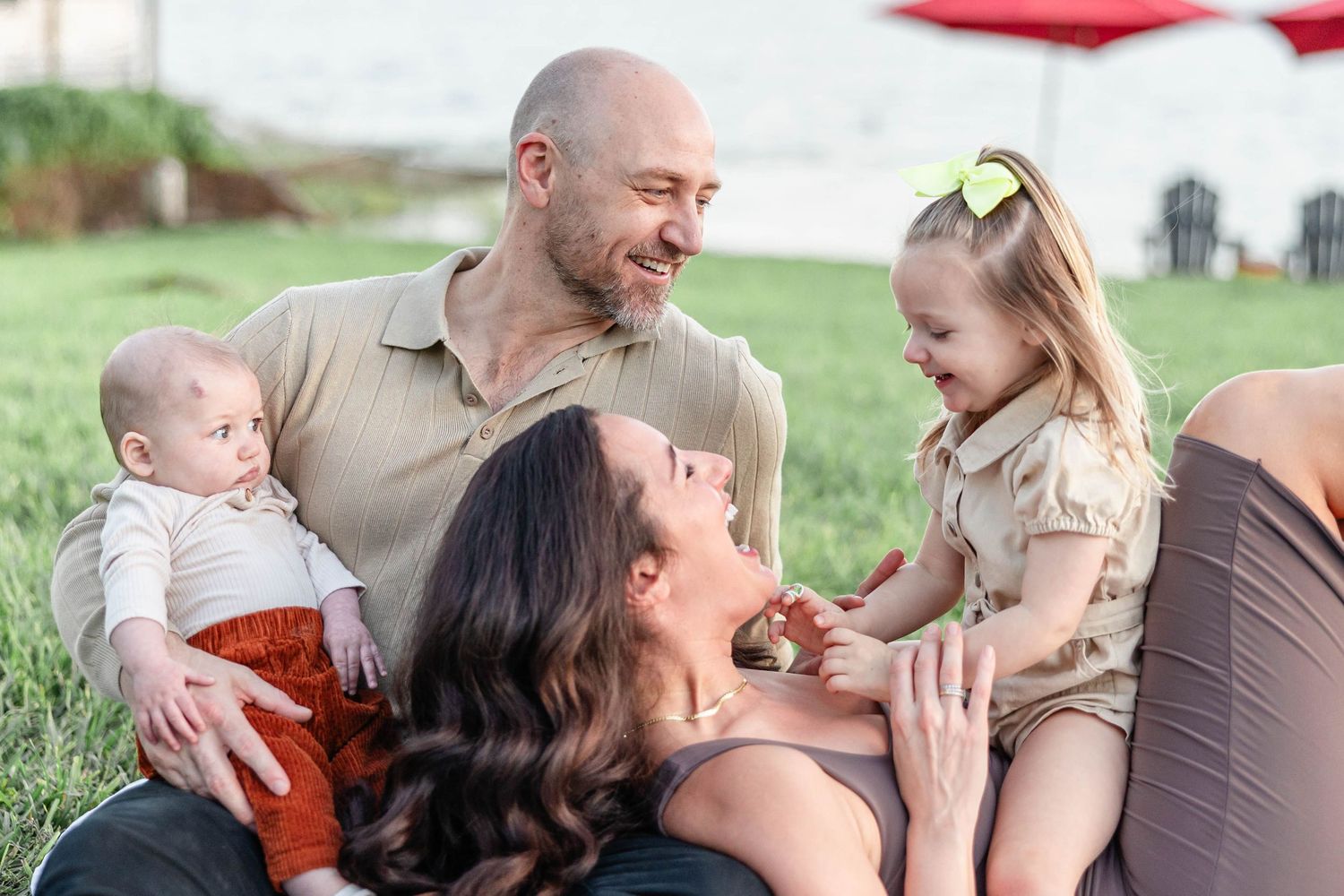 Happy family of four sitting on grass, parents smiling at toddler girl and baby outdoors.