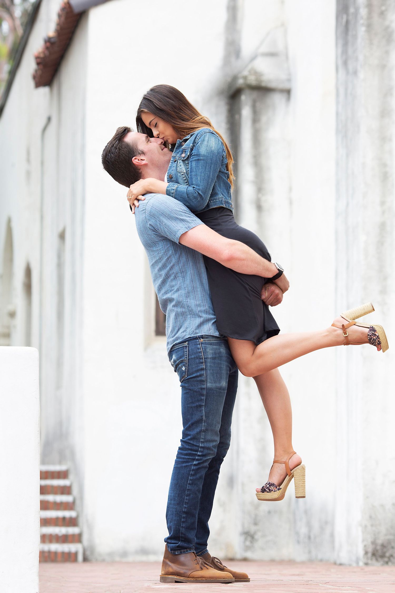 A man lifts and embraces a woman in heels near a white wall, both leaning in for a kiss.