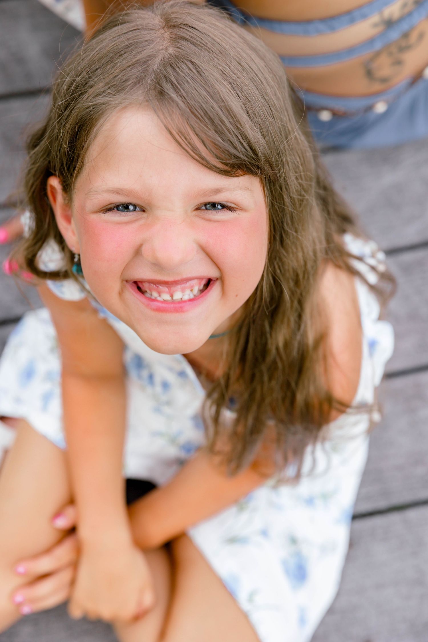 A young girl with long brown hair smiles broadly at the camera, sitting outdoors in a white floral top.