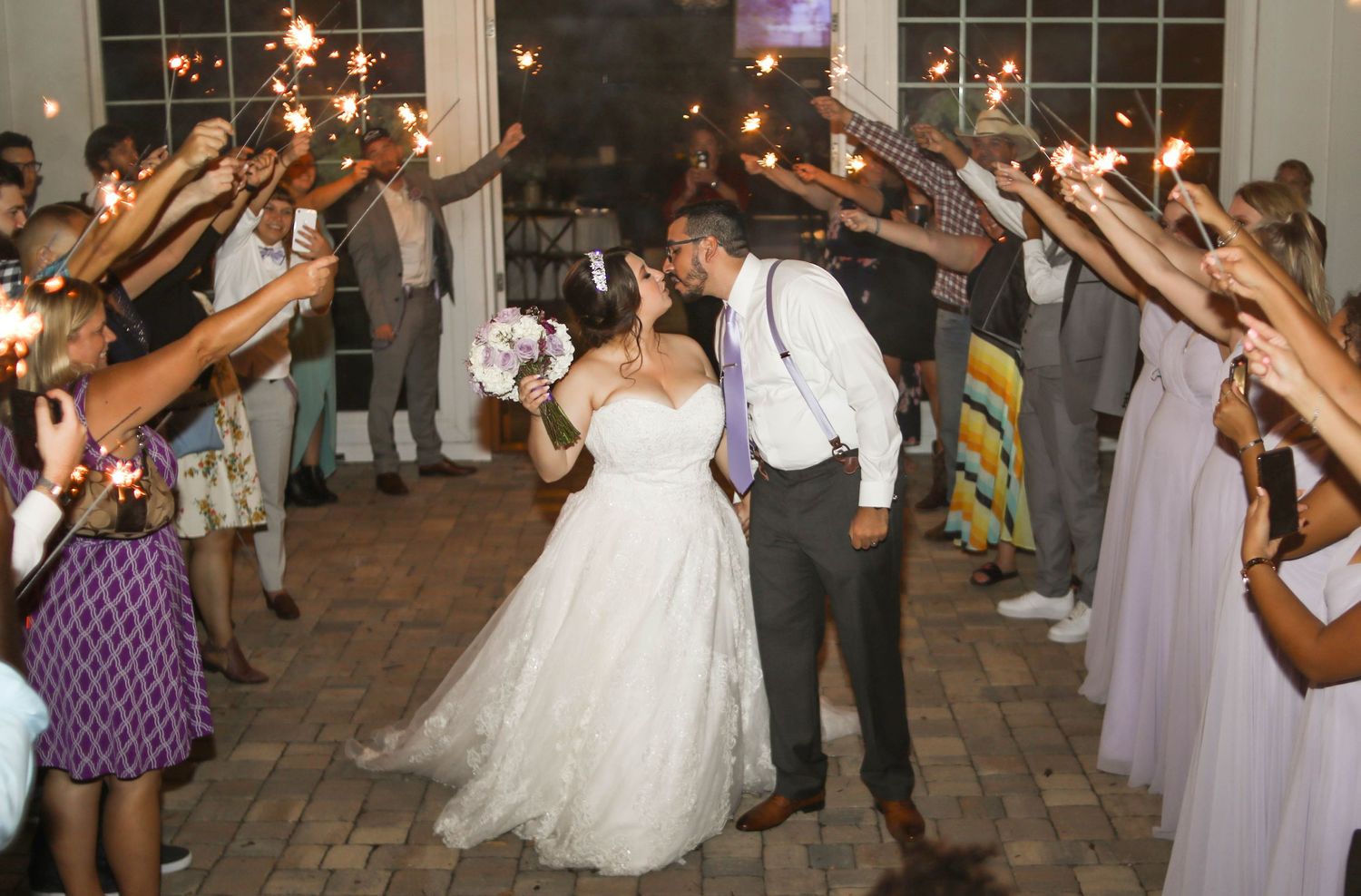 Newlyweds kiss during sparkler send-off exit surrounded by cheering wedding guests at night.