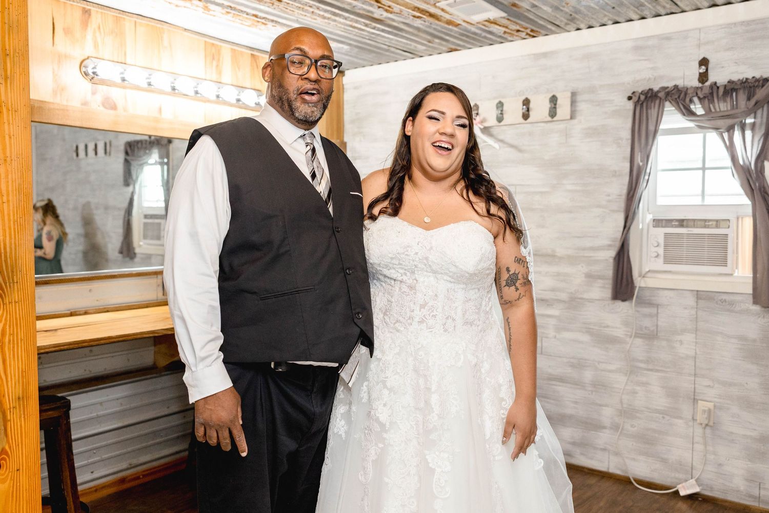 Bride in lace wedding gown and groom in vest smiling together inside a rustic wooden venue.