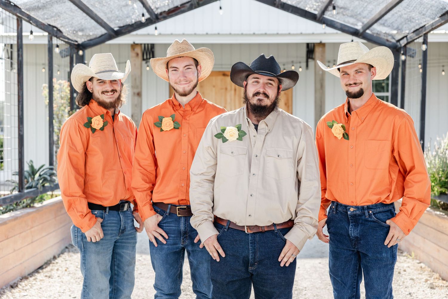 Groom and groomsmen in orange shirts, jeans, and cowboy hats posing in greenhouse.