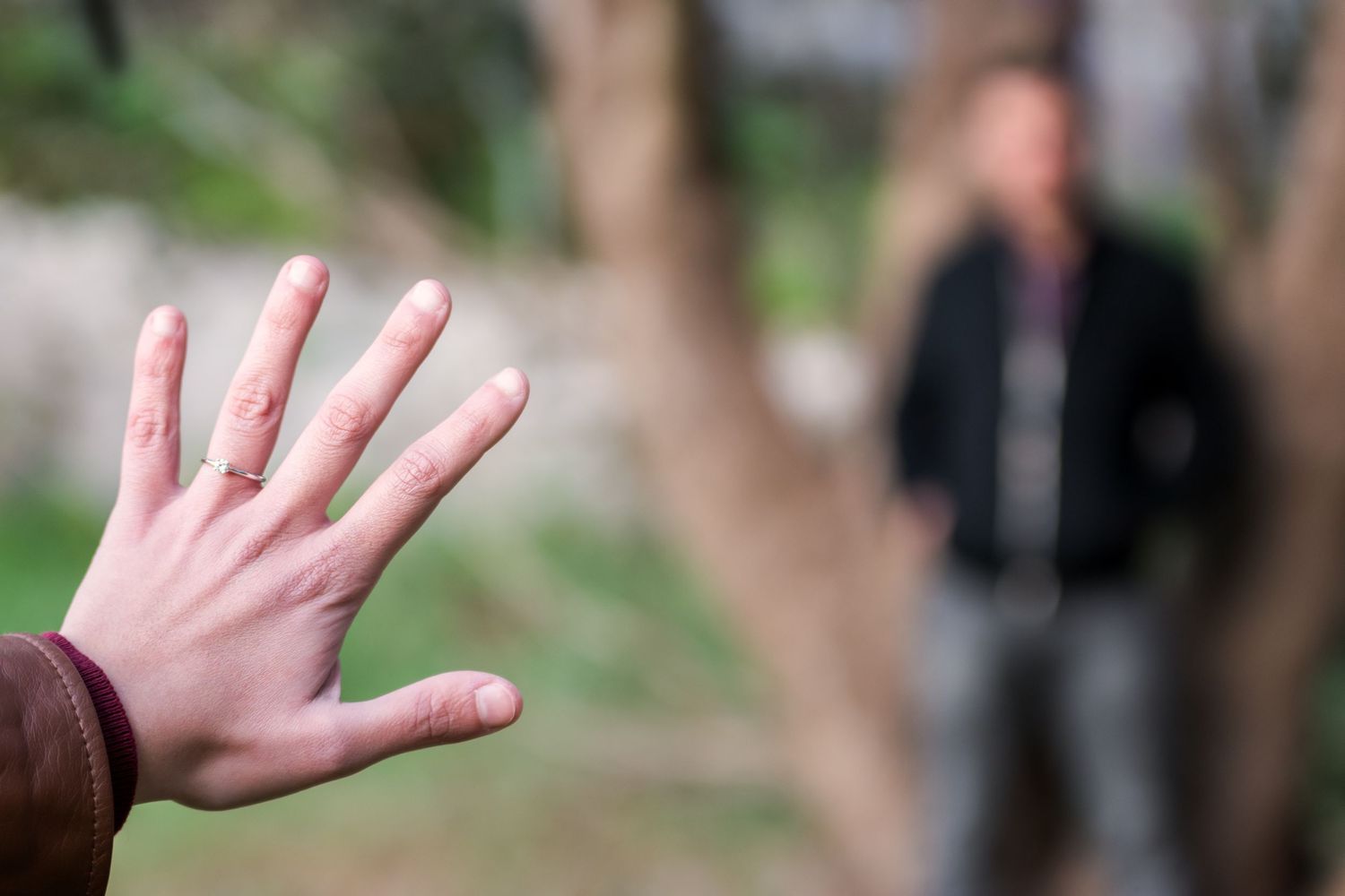 A hand reaches toward the camera in the foreground as a blurred figure stands on a park path behind.