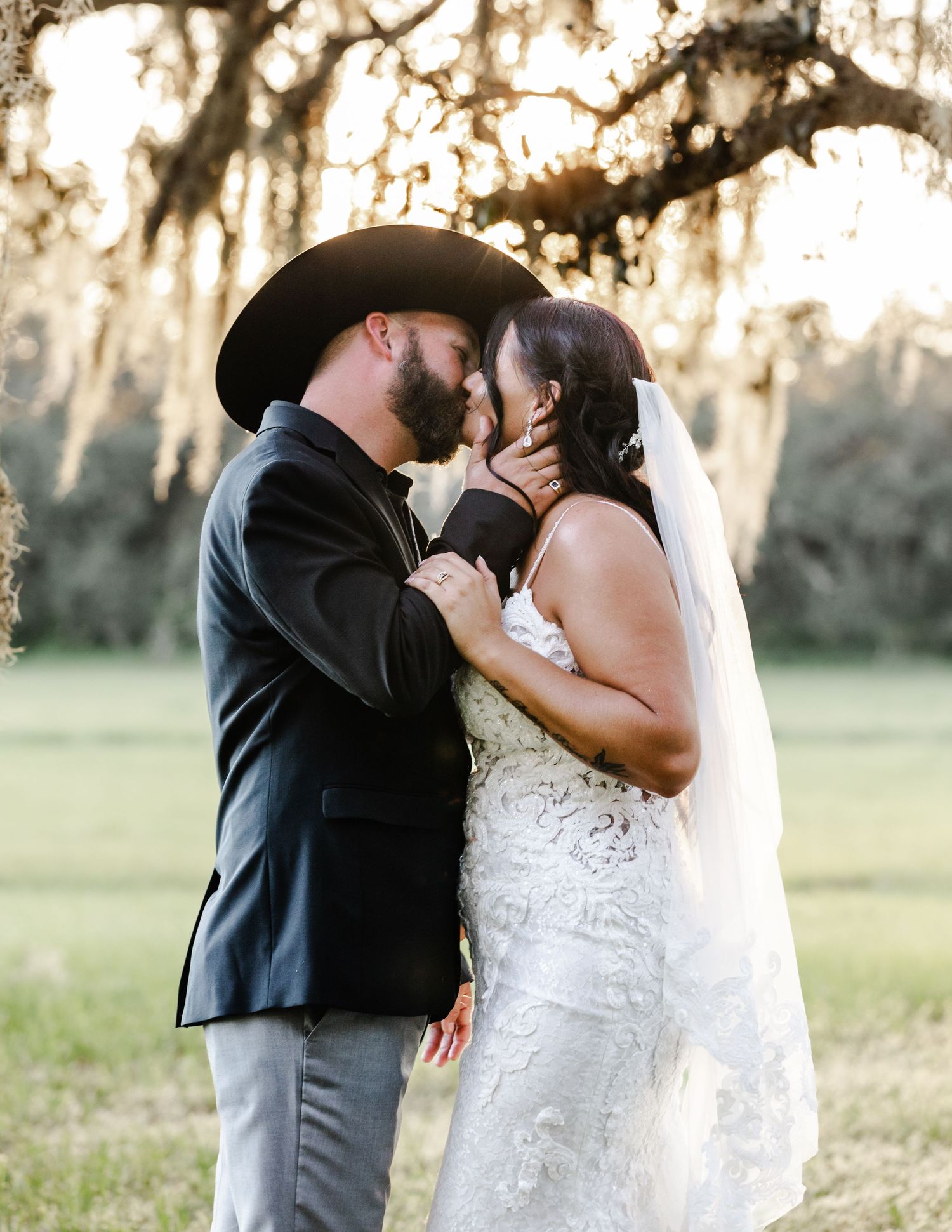 Groom in black cowboy hat and bride in lace gown share a romantic kiss under Spanish moss trees.