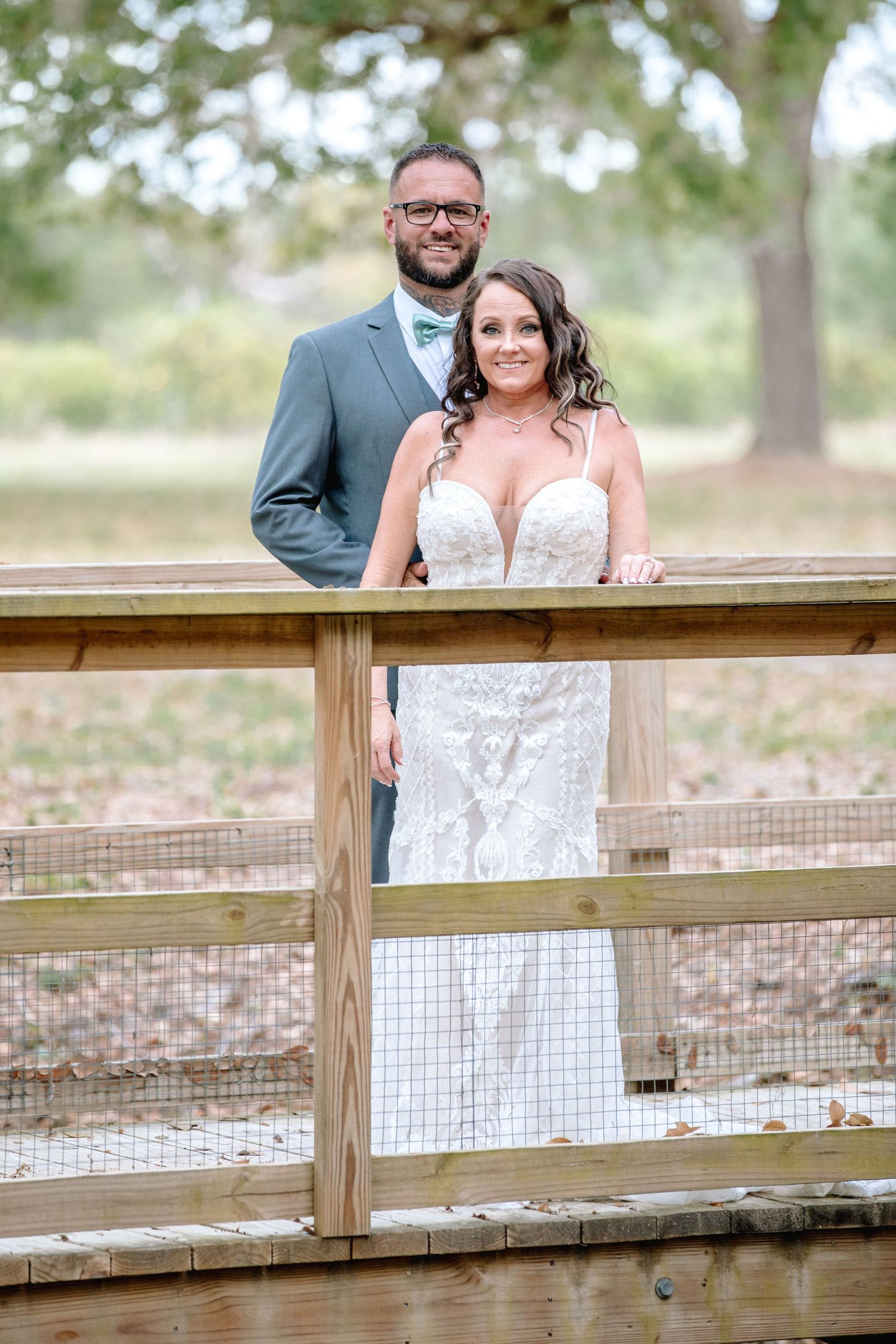 Bride and groom posing on a wooden bridge at an outdoor wedding venue surrounded by trees.
